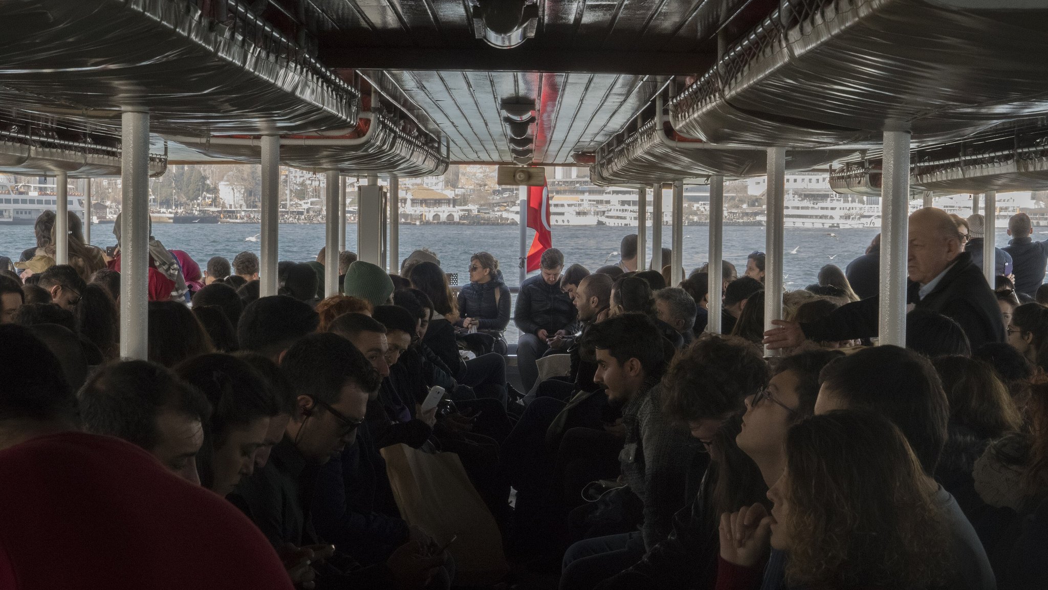 The packed lower deck of a Bosphorus ferry, passengers on the wooden benches under the canopy
