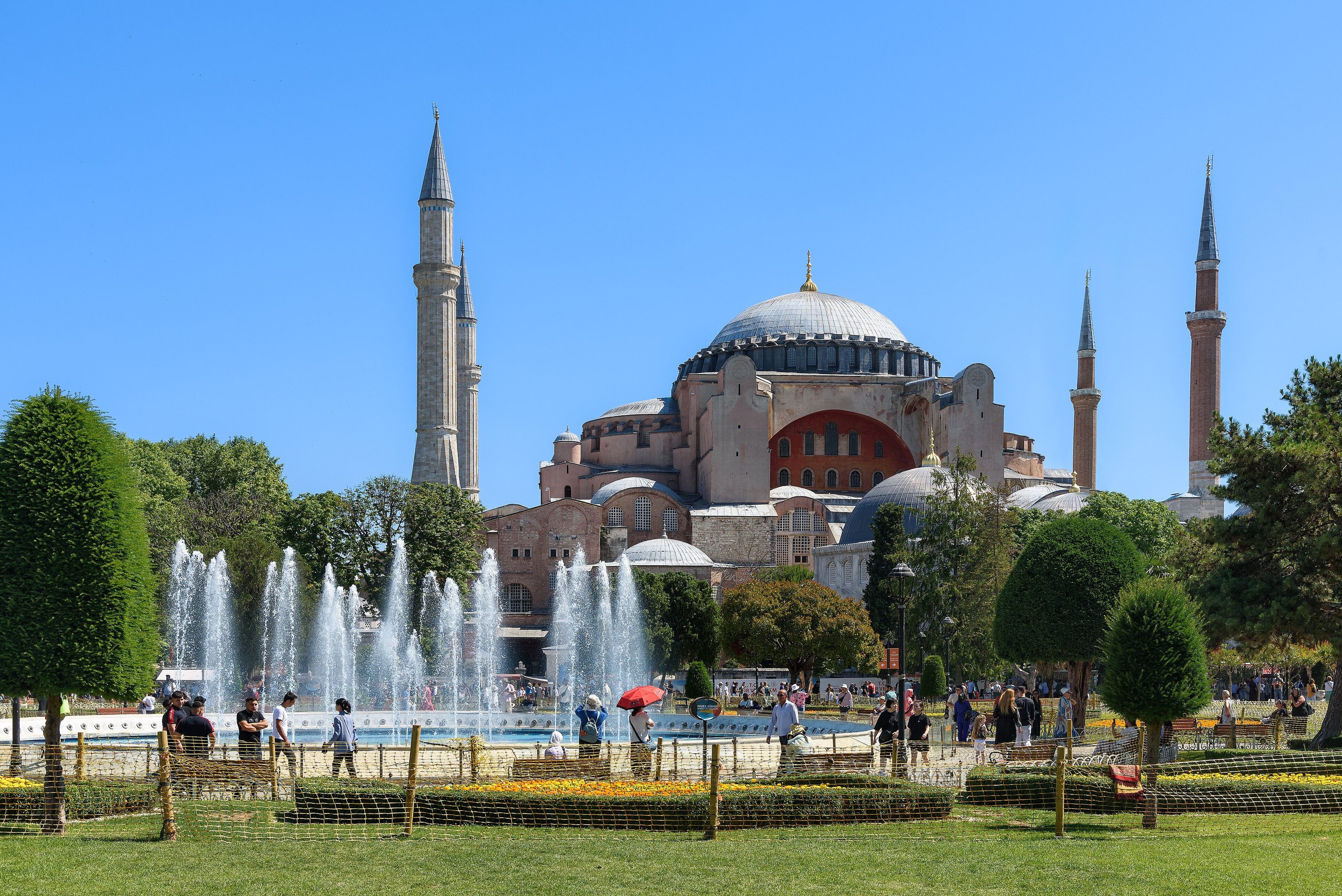 Hagia Sophia in full sun above the fountains of Sultanahmet park, its grey dome and four minarets against a deep blue sky, with people walking the paths in front