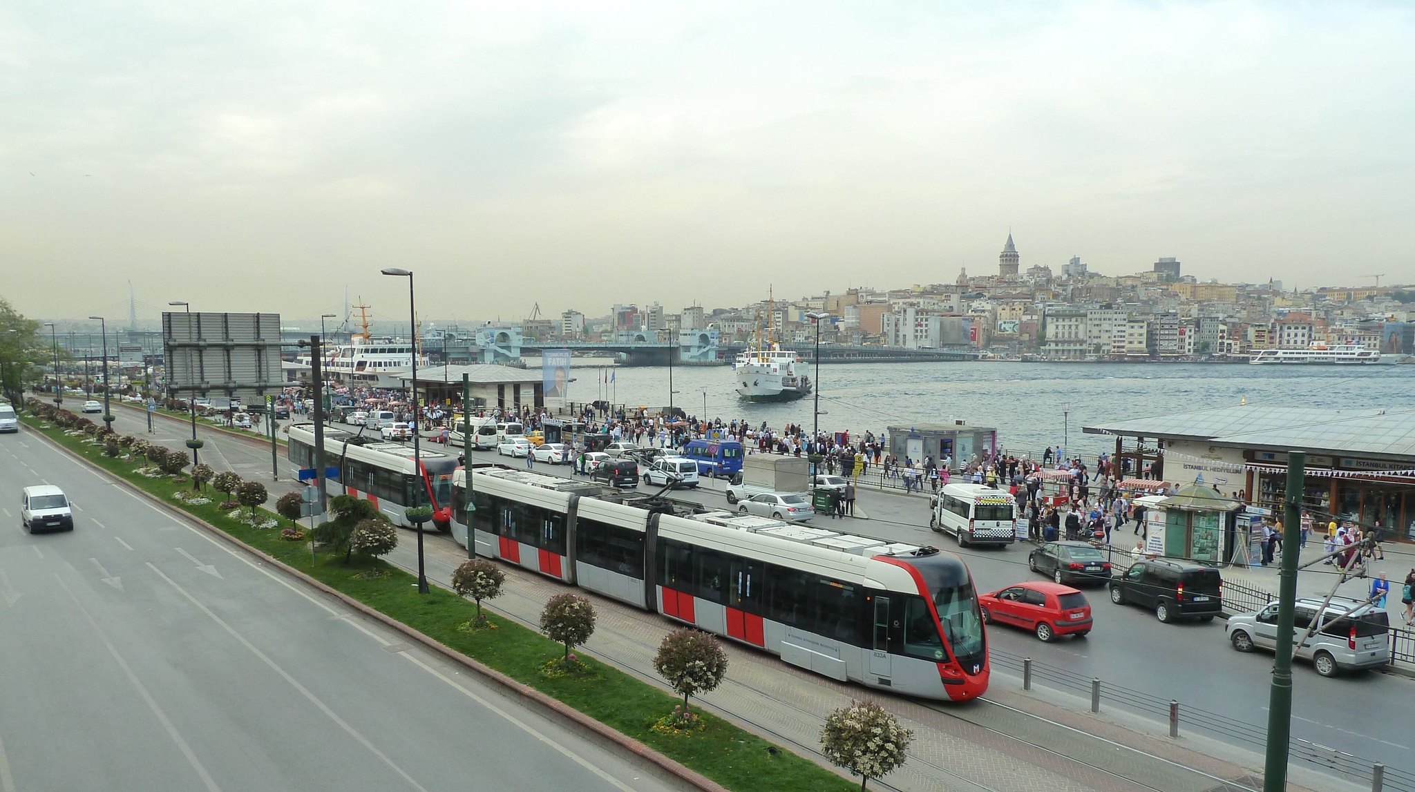 A red T1 tram running along the Eminonu waterfront with ferries at the piers and the Galata Tower on the skyline behind